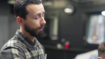 le coiffeur coupe les cheveux du client tout en parlant à quelqu'un hors caméra video