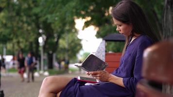 Young brunette woman sits on a park bench using smart phone and holding notebook video