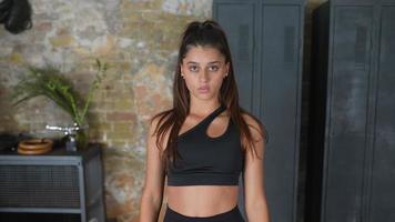 Athletic fit women in black sport bra faces and walks toward camera in room with lockers and brick walls video