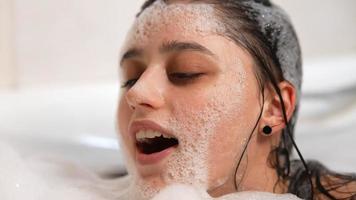 mujer joven en baño de burbujas se ríe y sopla espuma de las manos y acaricia burbujas en la cara video