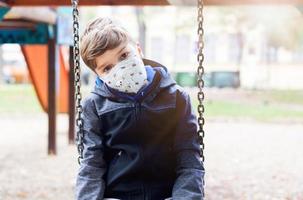 niño solitario con mascarilla sentado en un columpio en un patio vacío. foto