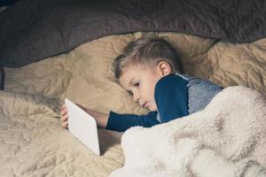 Tired little boy lying in bed and watching cartoons on touchpad. photo