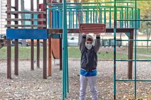 Small boy wearing protective face mask while playing with a swing on the playground.