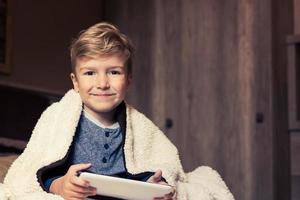 Smiling boy wrapped in blanket using touchpad in bedroom. photo
