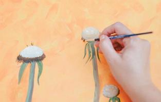 hand of adult woman painting with thin brush. orange canvas background. artwork of flowers in process photo