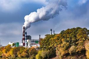 pipes of woodworking enterprise plant sawmill near river with autumn red yellow trees. Air pollution concept. Industrial landscape environmental pollution waste of thermal power plant photo