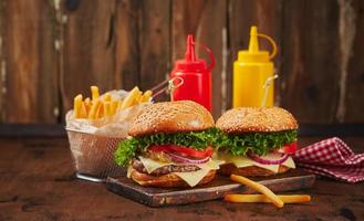 Two homemade burgers with beef, cheese and onion marmalade on a wooden board, fries in a metal basket and sauces. Fast food concept, american food photo