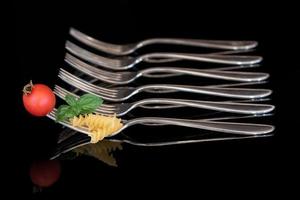 A large metal fork with pasta, basil and a tomato on it. In the background are empty forks in a row against a dark background. photo