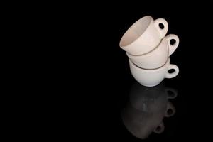 Three white, old espresso cups are stacked on the edge. The background is black. The cups are reflected on the ground. There is space for text. photo