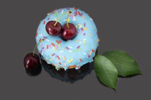 Close-up of a blue donought with red cherries reflected on a gray background. There are also cherry leaves to see. photo