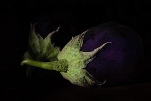 tiro oscuro de dos berenjenas moradas con tallos verdes que yacen uno al lado del otro sobre un fondo oscuro. foto