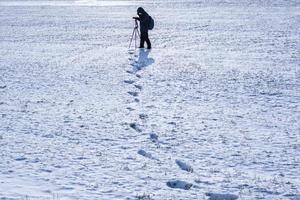 photographer with a tripod in snowy field takes pictures of landscape, footprints in snow photo