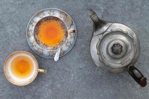 una vieja tetera de metal, una vieja taza de té con té y platillo y una vieja taza de té sin platillo se encuentran una al lado de la otra sobre un fondo gris. son fotografiados desde arriba. foto