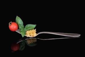A large metal fork with pasta, basil and a tomato on it. The cutlery is reflected against a dark background. photo