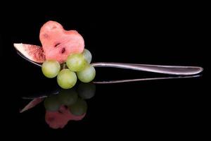 A piece of melon in the shape of a heart, fresh grapes and a piece of fig are placed on a metal spoon. The fruit salad is reflected on the dark background. photo