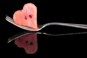 Close-up of a metal fork, with long tines, on which lies a piece of water belone in the shape of a heart. The fork is reflected on the dark background. photo