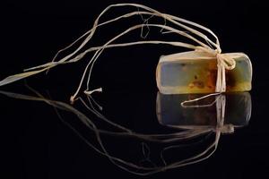 A piece of homemade soap is wrapped in raffia and reflected against a dark background photo