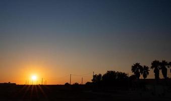 puesta de sol en sicilia. el cielo cambia de azul a naranja. brillan los rayos del sol. palmeras y otros árboles dan sombra entre los cables eléctricos y las torres. foto