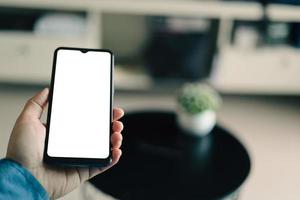 Mockup image of a man holding black mobile phone with blank white screen. In living room at home. photo