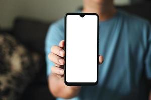 presentación de pantalla en blanco de teléfono inteligente. un joven sostiene un teléfono con una maqueta de pantalla blanca vacía. un hombre usa una camiseta azul en la sala de estar de su casa. foto