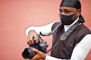 Young professional african american videographer holding professional camera with pro equipment. Afro cameraman wearing black duraq and face protect mask, making a videos. photo