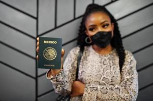 African american woman wearing black face mask show Mexico passport in hand. Coronavirus in America country, border closure and quarantine, virus outbreak concept. photo