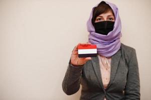 retrato de una joven musulmana que usa ropa formal, protege la máscara facial y el pañuelo de la cabeza del hiyab, sostiene la tarjeta de la bandera de yemen contra un fondo aislado. concepto de país coronavirus. foto