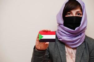 Portrait of young muslim woman wearing formal wear, protect face mask and hijab head scarf, hold Sudan flag card against isolated background. Coronavirus country concept. photo