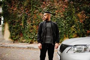 retrato de un elegante hombre de barba árabe con cuello alto gris y chaqueta negra. chico modelo árabe parado cerca de su coche. foto