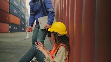 Young male Asian worker in safety uniform and hardhat rest from logistics work drinks water, talk and relax with female colleague at stacks of containers, shipping goods, and cargo transport industry. video