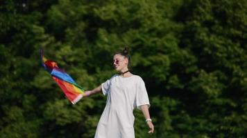 Young woman in white with sunglasses and top knots holds Pride flag and waves it in the wind in front of trees ata park video