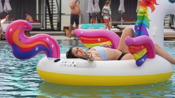 Young woman in a bikini rests on a big rainbow unicorn float laughing and reaching toward camera video