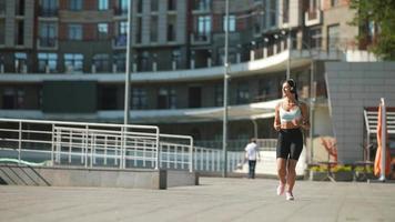 Fit young woman wearing headphones and sports bra and shorts runs along a concrete walkway in an urban space video