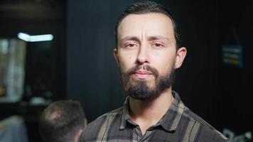 männlicher friseur steht vor der kamera, die im geschäft blickkontakt mit dem kunden im hintergrund herstellt video
