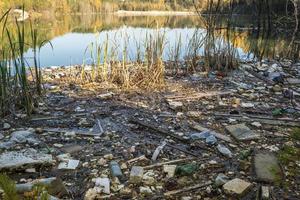 montones de residuos de construcción, residuos domésticos, espuma y botellas de plástico en la orilla de un lago forestal, problemas de contaminación ambiental foto