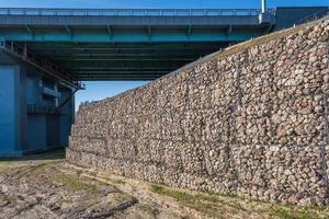 estructura de ingeniería hecha de piedras detrás de una malla metálica para fortalecer la orilla del río cerca del puente de la carretera foto