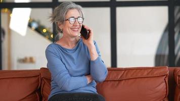 Woman with gray hair and glasses sits on a couch talking on a smart phone