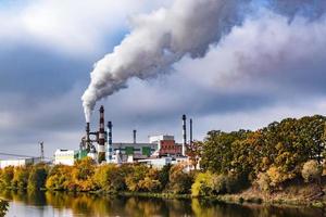 pipes of woodworking enterprise plant sawmill near river with autumn red yellow trees. Air pollution concept. Industrial landscape environmental pollution waste of thermal power plant photo