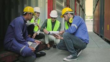 Group of multiracial workers people in safety uniforms and hardhats sit and discuss in stacks of logistics containers with male White manager, trading shipping goods for cargo transportation industry. video