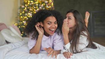 Two young women sit on bed in pajamas and talk with Christmas tree in background video