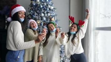 Junge Frauen auf einer Weihnachtsfeier mit festlichen Hüten tanzen und lachen mit Wunderkerzen vor einem Weihnachtsbaum video
