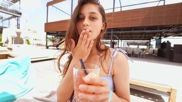 Young woman in swimsuit holds a drink by the pool tilts head and blows a kiss at the camera video