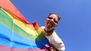 Young woman with sunglasses and top knots holds Pride flag and sticks out tongue  at low camera angle video