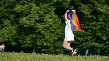 Young woman in white with sunglasses and top knots holds Pride flag and waves it in the wind in front of trees video