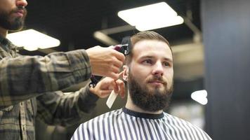 Le coiffeur coupe les cheveux d'un client masculin avec un peigne et une tondeuse video