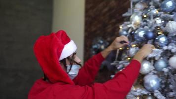 junger mann in rotem hemd und weihnachtsmütze schmückt einen weihnachtsbaum, während er eine maske trägt video
