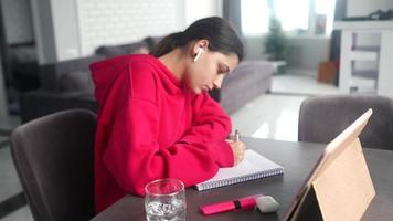 Young woman in a bright pink sweatshirt sits at a table with a tablet earbuds while writing in a notebook with a pen video