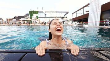 Young woman in a pool looks at and swims toward camera laughs and smiles while others lounge poolside video