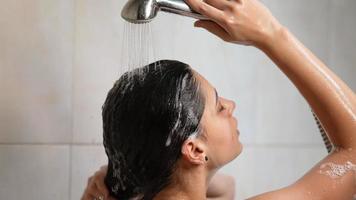 Woman uses handheld shower head to rinse shampoo from long dark hair in slow motion video