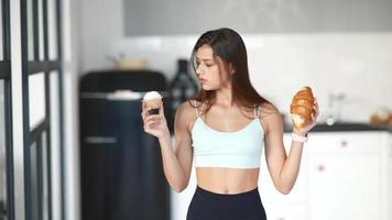 Young woman in active wear holds cupcake and croissant and studies both before taking a bite of the bread video